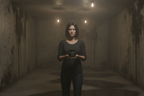 Woman holding a small object in a dimly lit room with peeling walls.